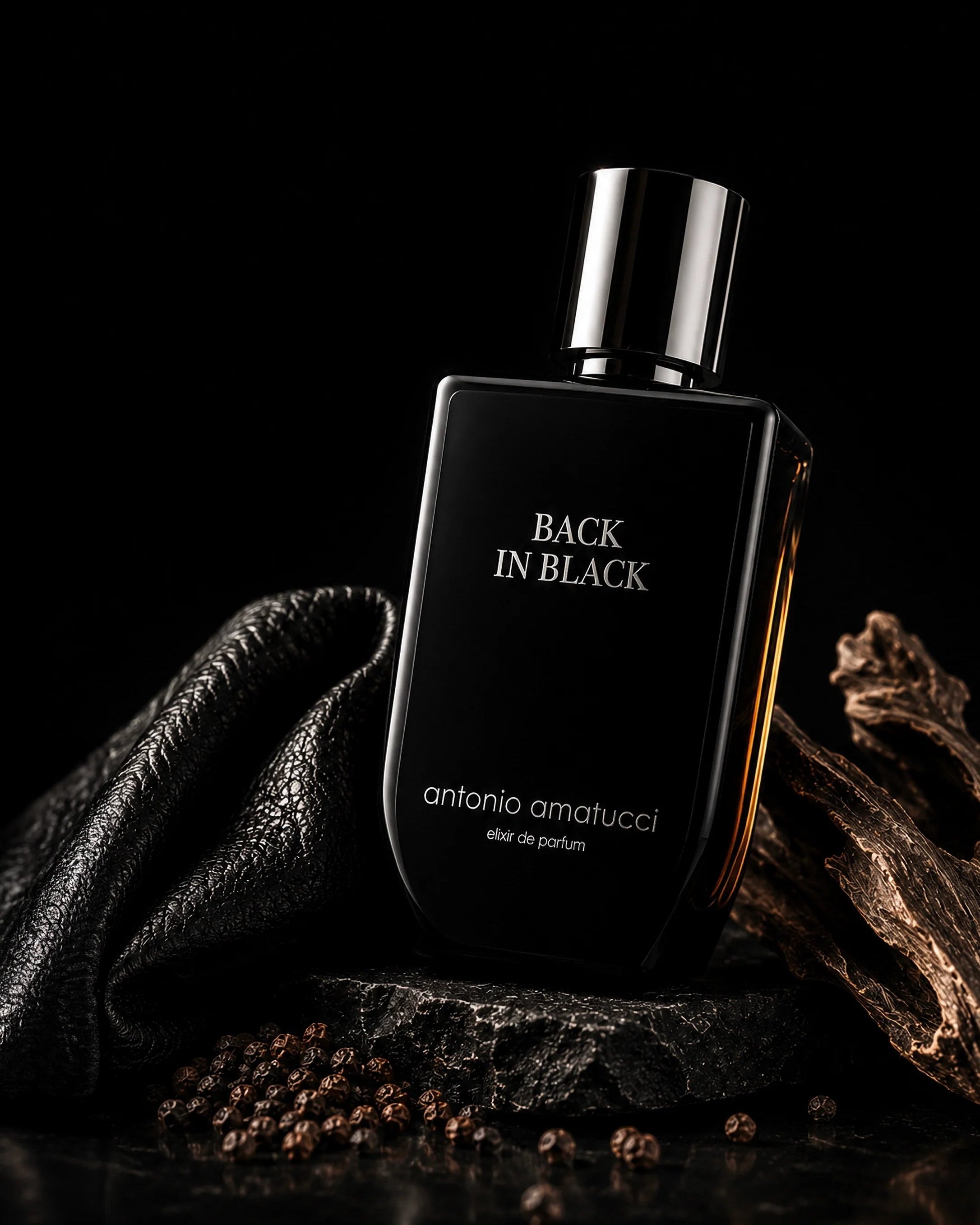 Back in Black di Antonio Amatucci. Il flacone in vetro nero lucido è adagiato su una pietra lavica scura, circondato da pelle nera martellata, rami di legno antico scuro e grani di pepe nero sparsi su una superficie lucida riflettente.