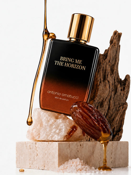 Bring Me The Horizon di Antonio Amatucci. La bottiglia di vetro sfumata dal nero al bronzo poggia su una roccia di sale rosa e un blocco di travertino, affiancata da un pezzo di legno antico e un dattero lucido da cui cola del miele dorato.