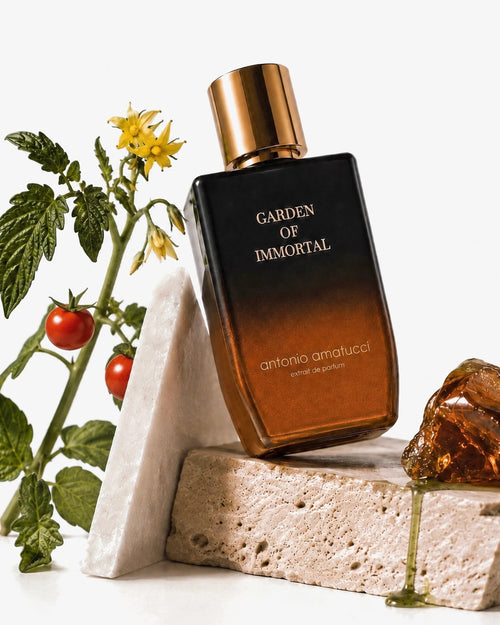 Garden of Immortal di Antonio Amatucci. Il flacone in vetro nero e sfumatura bronzo è adagiato su blocchi geometrici di marmo bianco e travertino. Accanto alla bottiglia si trovano una pianta con piccoli pomodori ciliegino e fiori gialli, e una resina ambrata lucida su cui cola un liquido verde.