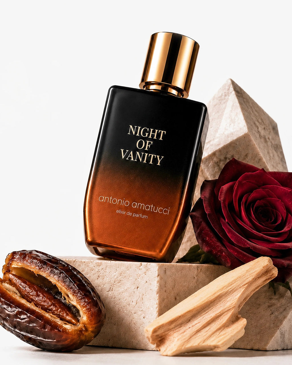 Night of Vanity di Antonio Amatucci. Il flacone in vetro con sfumatura nera e bronzo poggia inclinato su blocchi geometrici in pietra travertino, affiancato da una rosa rossa vellutata, un dattero aperto e un pezzo di legno chiaro aromatico.