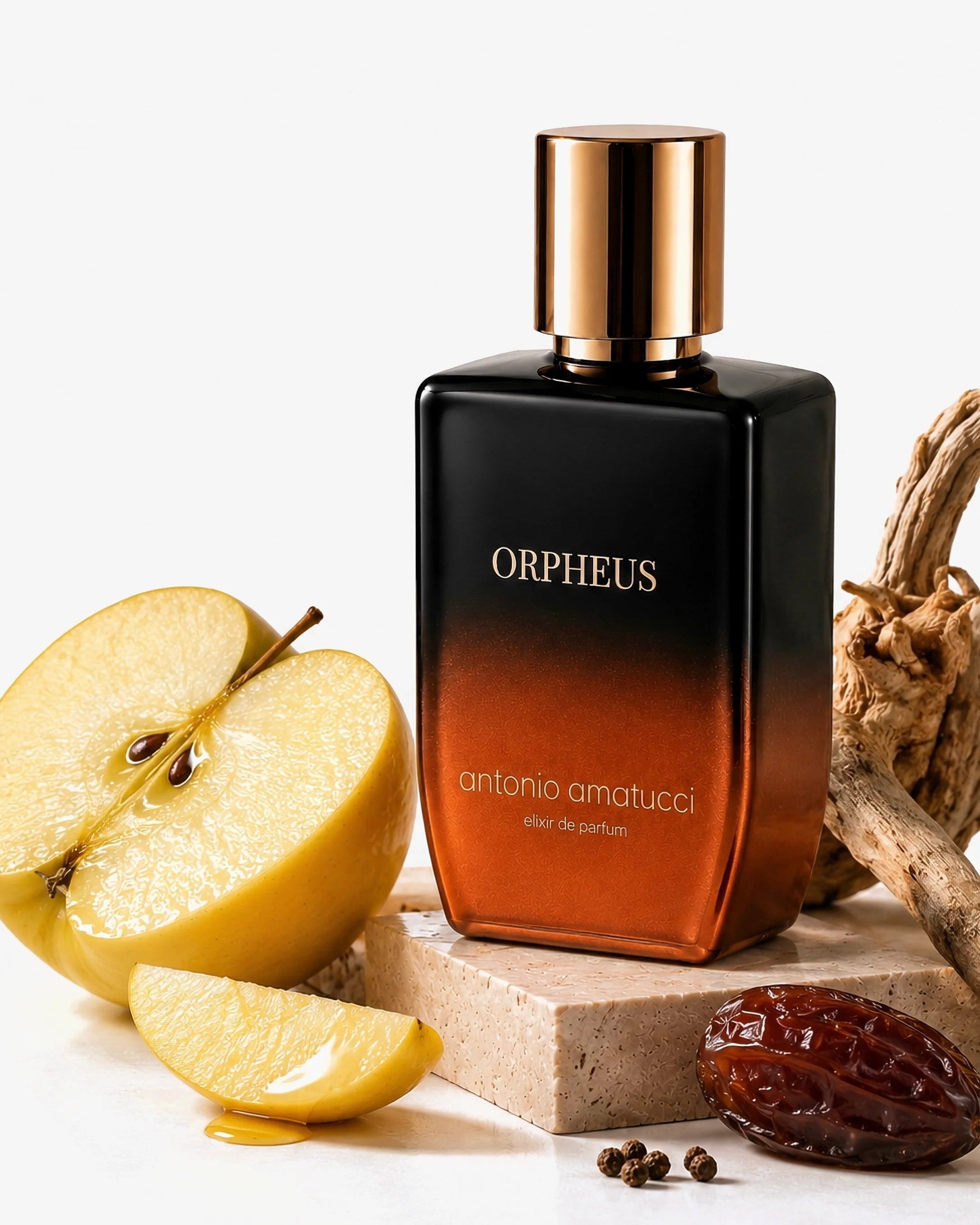 Orpheus di Antonio Amatucci. La bottiglia con sfumatura nera e bronzo è posizionata verticalmente su un blocco di travertino, circondata da una mela gialla tagliata a metà, uno spicchio di mela bagnato da una goccia dorata, un dattero lucido, grani di pepe nero e rami di legno antico ritorti.